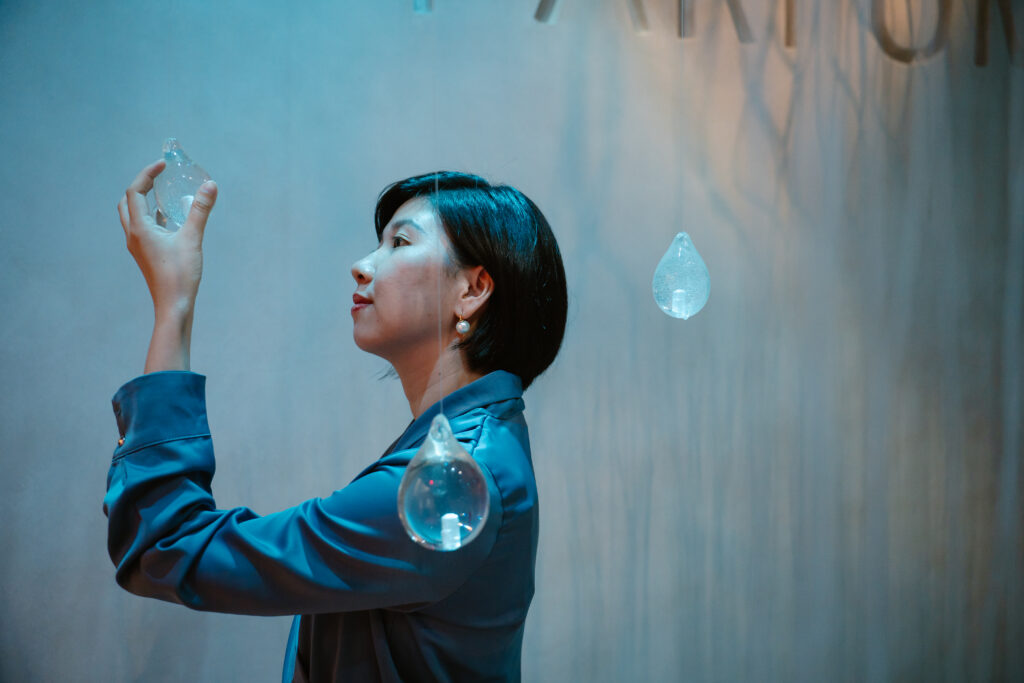 Woman in a blue shirt touches a hanging glass light bulb in a blue-toned installation. triển lãm mùi hương scent exhibition