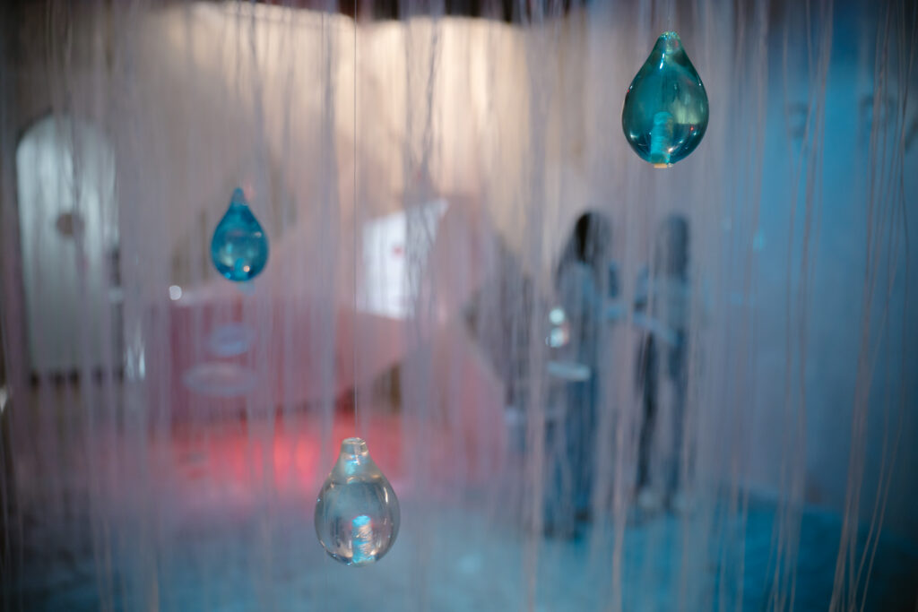 Blue glass teardrop ornaments hanging in a softly lit installation with blurred background hues of pink and blue. triển lãm mùi hương scent exhibition